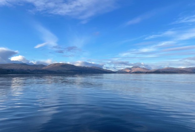 A photograph of Loch Lomond under a blue sky with white, wispy clouds